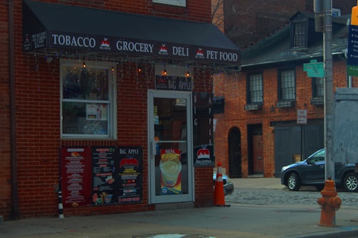A corner convenience store with signs advertising tobacco, grocery, deli, and pet food. The storefront features a black awning with white text, and there's a door with a menu and drink advertisements. Brick buildings and a parked car are visible in the background, with a prominent orange traffic cone and a fire hydrant nearby.