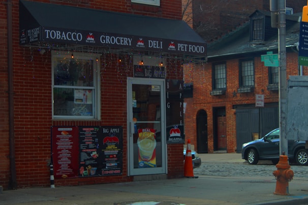 A corner convenience store with signs advertising tobacco, grocery, deli, and pet food. The storefront features a black awning with white text, and there's a door with a menu and drink advertisements. Brick buildings and a parked car are visible in the background, with a prominent orange traffic cone and a fire hydrant nearby.