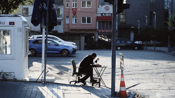 A person sits at a makeshift table on the side of a street, seemingly engaged in some activity like writing or reading. The surrounding area includes a parked blue car, a small building on the left with a sign, and a street with visible traffic cones. The buildings in the background have signs and apartments with visible windows. The overall scene appears urban and captures a moment of solitude in a busy environment.