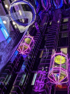 The image depicts a futuristic interior view with vibrant neon lighting. The walls are covered in reflective, dark-colored tiles, interspersed with bright neon lights in geometric patterns. There are two prominent, illuminated structures resembling lanterns or futuristic elevators, adorned with intricate designs and glowing in neon purple. The ceiling features an intricate arrangement of illuminated circular shapes, adding to the sci-fi ambiance.