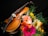 A violin rests on a black surface surrounded by a vibrant bouquet of gerbera daisies in various colors, including pink, orange, yellow, and white. Green foliage complements the flowers and contrasts with the dark background, highlighting the polished wood and strings of the violin.