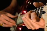 A close-up of hands playing a guitar, focusing on the fingers pressing down on the strings and the frets. The guitar neck is prominent with visible strings and frets, while the background is softly blurred. The lighting highlights the fingertips and the glossy finish of the instrument.