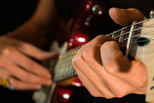 A close-up of hands playing a guitar, focusing on the fingers pressing down on the strings and the frets. The guitar neck is prominent with visible strings and frets, while the background is softly blurred. The lighting highlights the fingertips and the glossy finish of the instrument.