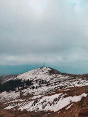 A mountainous landscape with patches of snow covering the ground, surrounded by a range of bare trees. A communication tower stands on the hilltop under an overcast, cloudy sky.