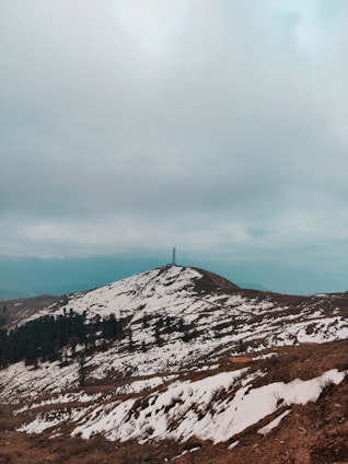 A mountainous landscape with patches of snow covering the ground, surrounded by a range of bare trees. A communication tower stands on the hilltop under an overcast, cloudy sky.