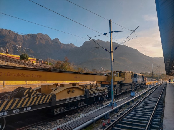 A train with a large breakdown crane attached is stationed at a railway platform. The backdrop features towering mountains under a clear blue sky. The tracks extend into the distance, and the platform is partially visible on the right side. There are a few small figures further down the platform.