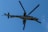 A military helicopter with large rotating rotor blades is flying against a clear blue sky. The helicopter has a camouflage paint scheme and appears to have armament mounted on it.