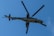 A military helicopter with large rotating rotor blades is flying against a clear blue sky. The helicopter has a camouflage paint scheme and appears to have armament mounted on it.