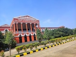 A large, classical-style red building featuring tall columns and a prominent triangular pediment at the top. The structure displays the words 'High Court of Karnataka Kalaburagi Bench' and is surrounded by lush greenery, including trees and bushes. The sky is clear and blue, and there is a paved pathway lined with yellow and black curbs leading to the building.