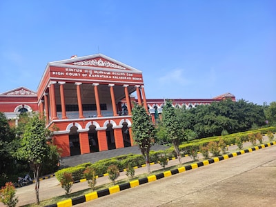 A large, classical-style red building featuring tall columns and a prominent triangular pediment at the top. The structure displays the words 'High Court of Karnataka Kalaburagi Bench' and is surrounded by lush greenery, including trees and bushes. The sky is clear and blue, and there is a paved pathway lined with yellow and black curbs leading to the building.