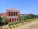 A large, classical-style red building featuring tall columns and a prominent triangular pediment at the top. The structure displays the words 'High Court of Karnataka Kalaburagi Bench' and is surrounded by lush greenery, including trees and bushes. The sky is clear and blue, and there is a paved pathway lined with yellow and black curbs leading to the building.