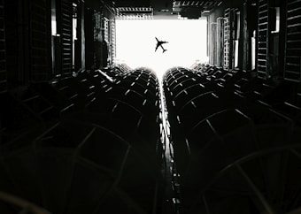 An airplane is silhouetted against a bright, overexposed sky seen through the inside of a dimly lit cargo plane. The interior features structural elements and rows of cylindrical shapes on both sides, leading the eye towards the opening where the airplane is visible.