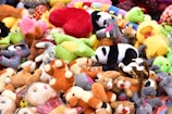A colorful assortment of plush toys piled together, featuring a panda, various animals, and heart-shaped objects. The toys are arranged in a chaotic yet vibrant manner, showcasing a diverse mix of colors and shapes.