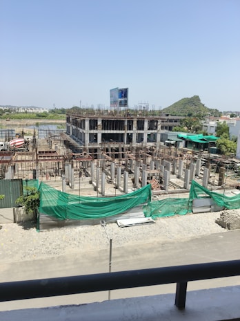 A construction site with a partially built multi-story building. The structure is made of concrete and appears to have several floors with scaffolding and metal reinforcement rods protruding from the incomplete sections. Green netting is draped over parts of the construction area to contain debris. A construction vehicle, specifically a cement mixer, is visible in the background. The surroundings include some vegetation and a distant hill under a clear blue sky.