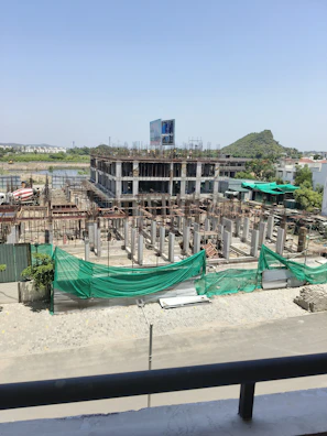 A construction site with a partially built multi-story building. The structure is made of concrete and appears to have several floors with scaffolding and metal reinforcement rods protruding from the incomplete sections. Green netting is draped over parts of the construction area to contain debris. A construction vehicle, specifically a cement mixer, is visible in the background. The surroundings include some vegetation and a distant hill under a clear blue sky.