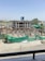 A construction site with a partially built multi-story building. The structure is made of concrete and appears to have several floors with scaffolding and metal reinforcement rods protruding from the incomplete sections. Green netting is draped over parts of the construction area to contain debris. A construction vehicle, specifically a cement mixer, is visible in the background. The surroundings include some vegetation and a distant hill under a clear blue sky.