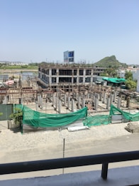 A construction site with a partially built multi-story building. The structure is made of concrete and appears to have several floors with scaffolding and metal reinforcement rods protruding from the incomplete sections. Green netting is draped over parts of the construction area to contain debris. A construction vehicle, specifically a cement mixer, is visible in the background. The surroundings include some vegetation and a distant hill under a clear blue sky.