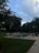 A serene backyard scene featuring a rectangular swimming pool surrounded by lush green grass and dense trees. There are several white lounge chairs lined up neatly beside the pool. The sky is partly cloudy with patches of blue visible, suggesting late afternoon or early evening. On the right, a patio area with outdoor furniture overlooks the pool, providing a comfortable relaxation spot.