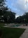 A serene backyard scene featuring a rectangular swimming pool surrounded by lush green grass and dense trees. There are several white lounge chairs lined up neatly beside the pool. The sky is partly cloudy with patches of blue visible, suggesting late afternoon or early evening. On the right, a patio area with outdoor furniture overlooks the pool, providing a comfortable relaxation spot.