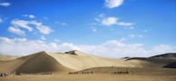 A vast desert landscape with large sand dunes under a bright blue sky. In the foreground, a caravan of camels is led by people, creating a line silhouetted against the desert. The sky is dotted with white clouds, and the light casts gentle shadows on the dunes.