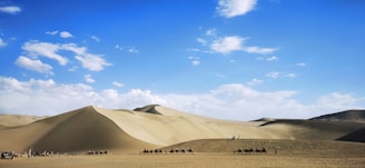 A vast desert landscape with large sand dunes under a bright blue sky. In the foreground, a caravan of camels is led by people, creating a line silhouetted against the desert. The sky is dotted with white clouds, and the light casts gentle shadows on the dunes.