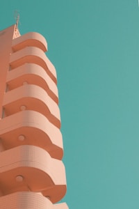 A building with a unique architectural design featuring curved balconies and a pale, pastel pink color stands against a clear turquoise sky. The structure exhibits a modern and minimalist aesthetic, with small round light fixtures under each balcony. The image conveys a sense of tranquility and simplicity through its clean lines and soft colors.