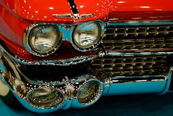A close-up view of a vintage car grille adorned with chrome detailing. The bright red color of the car's body complements the shiny metallic surface. Headlights are prominently featured, showcasing a classic design.