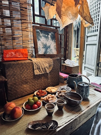 A rustic wooden table is set with a variety of food items, including fruits like peaches, green citrus, and red tomatoes, alongside dry snacks such as cookies and biscuits. Several ceramic cups and a teapot are placed on the table. The background includes a woven bamboo screen, a piece of art leaning against it, and paper decorations above. A warm, inviting atmosphere is created by the soft lighting and traditional materials.