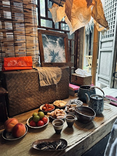A rustic wooden table is set with a variety of food items, including fruits like peaches, green citrus, and red tomatoes, alongside dry snacks such as cookies and biscuits. Several ceramic cups and a teapot are placed on the table. The background includes a woven bamboo screen, a piece of art leaning against it, and paper decorations above. A warm, inviting atmosphere is created by the soft lighting and traditional materials.