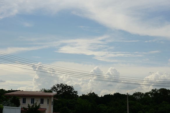 A residential house with a red roof is situated among lush green trees. Overhead utility lines stretch horizontally across the image against a partly cloudy sky with large billowing clouds. There is a mix of trees and foliage in the foreground.