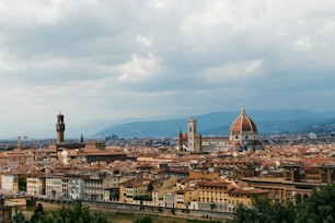 A panoramic view of an historic city with a prominent cathedral featuring a large dome. Numerous older buildings with terracotta rooftops are visible, set against a backdrop of verdant hills under a partly cloudy sky.