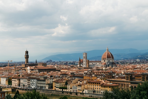 A panoramic view of an historic city with a prominent cathedral featuring a large dome. Numerous older buildings with terracotta rooftops are visible, set against a backdrop of verdant hills under a partly cloudy sky.