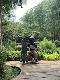 A person wearing a black helmet and dark clothing is crouching on a wooden pathway, holding an electric skateboard vertically. Lush green vegetation and tall trees surround the area, creating a serene natural setting.