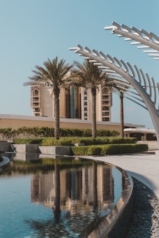 A modern architectural landscape featuring a luxurious building with tall palm trees in the foreground. There is also a reflective water feature creating a mirror image of the surroundings. The sky is clear and blue, enhancing the calm and serene atmosphere.