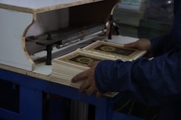 A person is handling a stack of books with ornate covers, situated on a blue table. The setting suggests a printing or binding process, and the person appears to be tending to a machine.