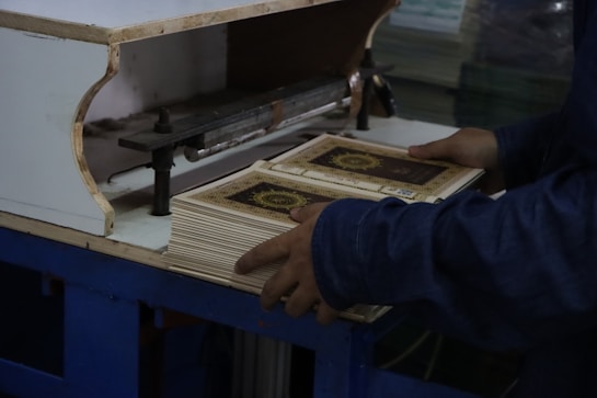 A person is handling a stack of books with ornate covers, situated on a blue table. The setting suggests a printing or binding process, and the person appears to be tending to a machine.