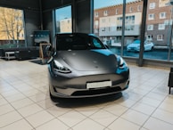 A sleek, modern electric car is displayed inside a showroom with large windows. The car has a futuristic design with smooth curves and a metallic finish, situated on a polished tiled floor. Large windows behind the car reveal views of the outside, including buildings and another car parked by the road.