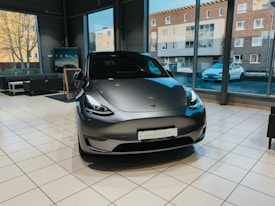 A sleek, modern electric car is displayed inside a showroom with large windows. The car has a futuristic design with smooth curves and a metallic finish, situated on a polished tiled floor. Large windows behind the car reveal views of the outside, including buildings and another car parked by the road.