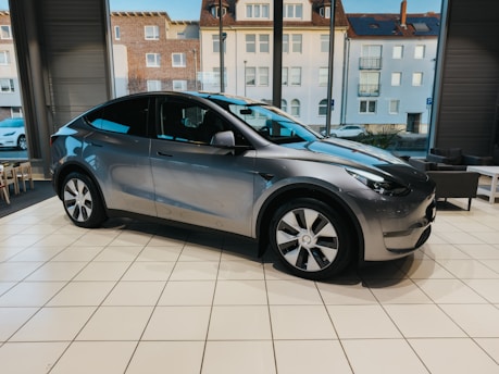 A sleek, modern electric car is displayed in a showroom with large windows showing an urban street view. The vehicle has a metallic gray finish and distinctive alloy wheels. The interior of the showroom is bright, with several chairs visible near the windows.