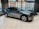 A sleek, modern electric car is displayed in a showroom with large windows showing an urban street view. The vehicle has a metallic gray finish and distinctive alloy wheels. The interior of the showroom is bright, with several chairs visible near the windows.