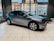 A sleek, modern electric car is displayed in a showroom with large windows showing an urban street view. The vehicle has a metallic gray finish and distinctive alloy wheels. The interior of the showroom is bright, with several chairs visible near the windows.
