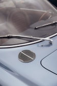 A close-up view of a vintage car's windshield, featuring two chrome wipers and a round grille on the blue hood. The car's interior, including brown seats, is visible through the windshield.