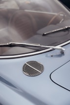 A close-up view of a vintage car's windshield, featuring two chrome wipers and a round grille on the blue hood. The car's interior, including brown seats, is visible through the windshield.