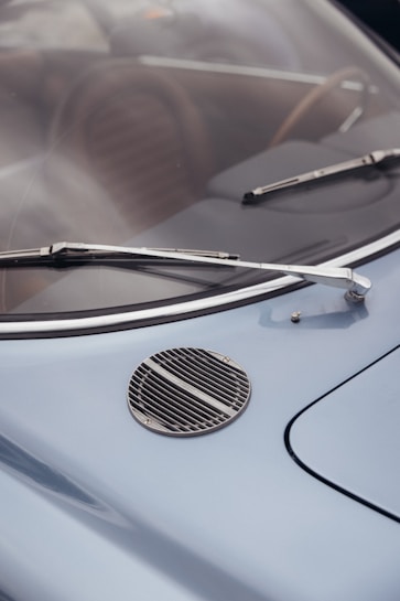 A close-up view of a vintage car's windshield, featuring two chrome wipers and a round grille on the blue hood. The car's interior, including brown seats, is visible through the windshield.