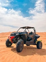 A rugged off-road utility vehicle with large, robust tires is parked on an expansive desert landscape. The red and black vehicle, with a roll cage and high ground clearance, stands prominently against the backdrop of vast sandy dunes under a bright, partly cloudy sky.