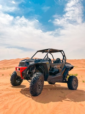 A rugged off-road utility vehicle with large, robust tires is parked on an expansive desert landscape. The red and black vehicle, with a roll cage and high ground clearance, stands prominently against the backdrop of vast sandy dunes under a bright, partly cloudy sky.