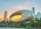 An architectural marvel featuring a futuristic building with an oval, hollow ring structure adorned with Arabic calligraphy. The background includes modern skyscrapers, palm trees, and a vivid sunset sky that creates a soft gradient of pastel colors.