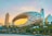 An architectural marvel featuring a futuristic building with an oval, hollow ring structure adorned with Arabic calligraphy. The background includes modern skyscrapers, palm trees, and a vivid sunset sky that creates a soft gradient of pastel colors.
