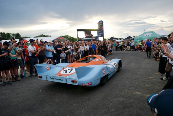 A classic racing car with blue and orange Gulf livery is surrounded by a large crowd of spectators. The scene appears to be an outdoor event, possibly a car show or race. People are taking photos and observing the car with interest. Tents and banners are visible in the background, indicating a festive or organized event.