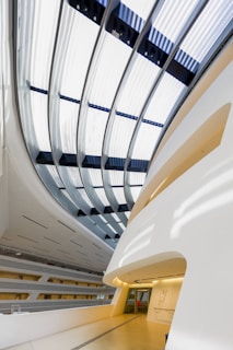 A modern architectural interior with sweeping, curved lines and a large, glass-paneled ceiling that allows natural light to flood the space. The walls and surfaces are primarily white and gray, creating a sleek and clean atmosphere. A corridor with a polished floor leads deeper into the structure.
